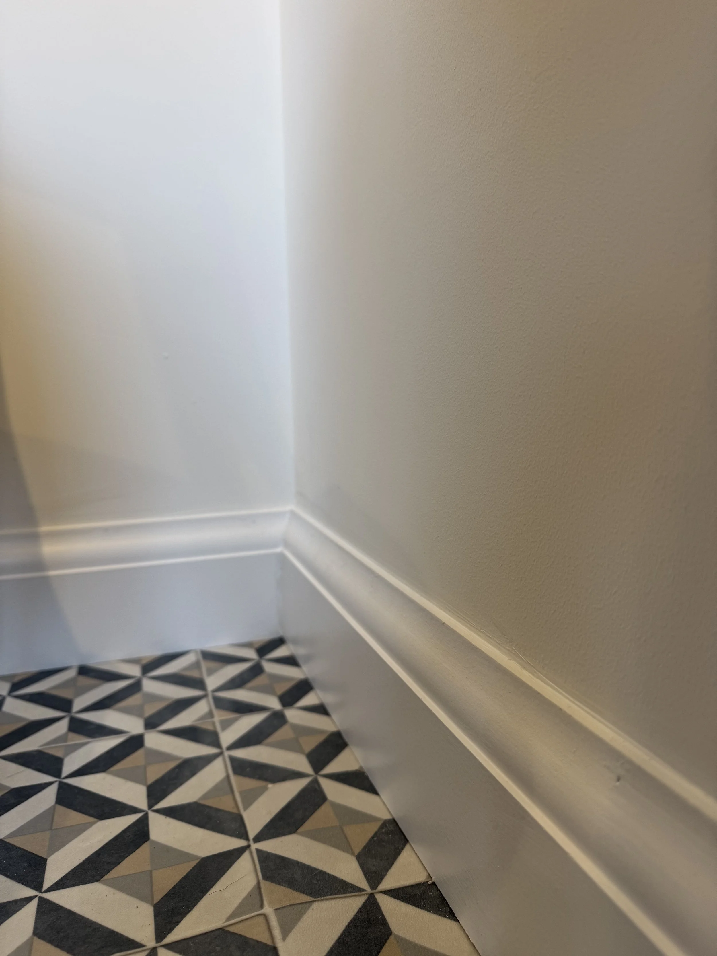 Close-up of a wall corner with patterned tile flooring and a white baseboard, showing the intersection of the wall and floor.