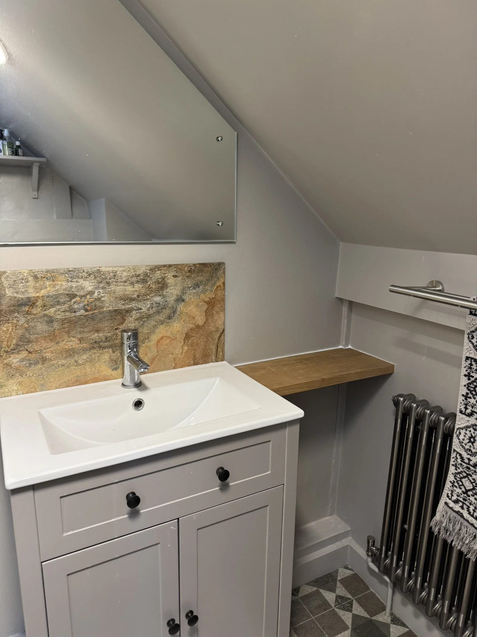 Small bathroom with a white vanity sink, a rectangular mirror, a slate backsplash, a wooden shelf, a patterned towel, and a black radiator.