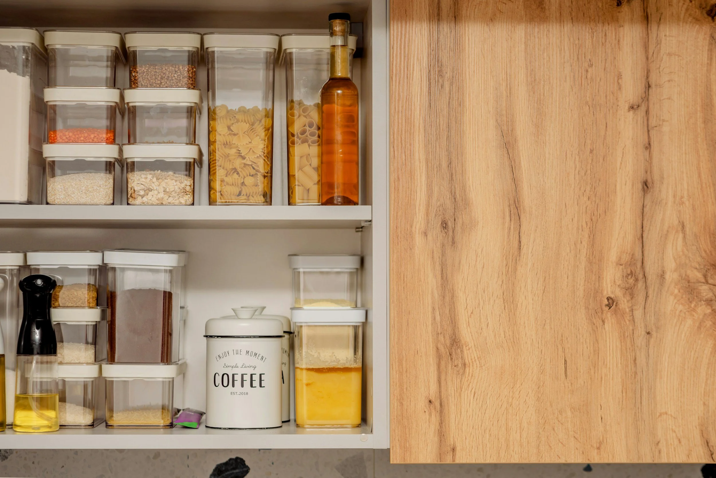Kitchen pantry with clear plastic containers filled with pasta, spices, grains, and other dry ingredients, a white coffee container with black text, a small jar with yellow liquid, and a wooden cabinet door on the right.