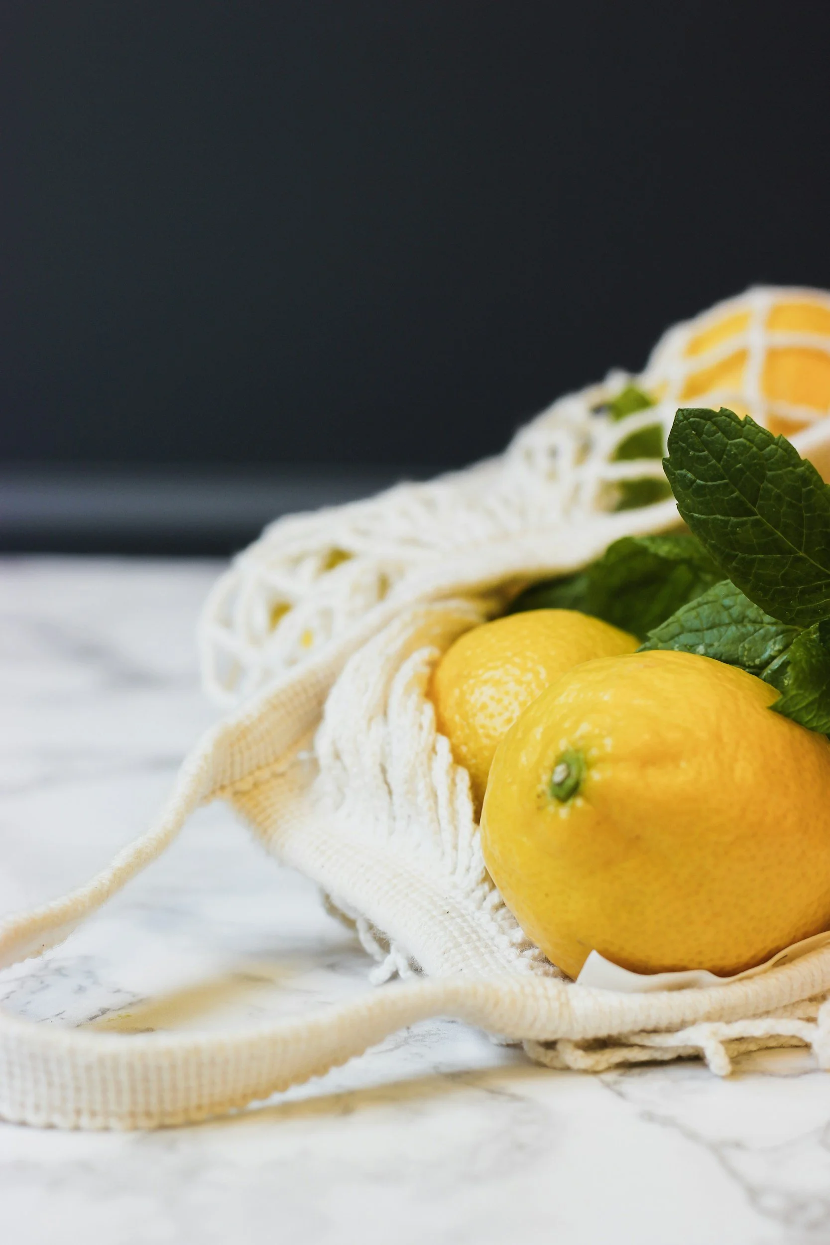 Yellow lemons with green mint leaves inside a reusable cloth bag on a white marble surface.
