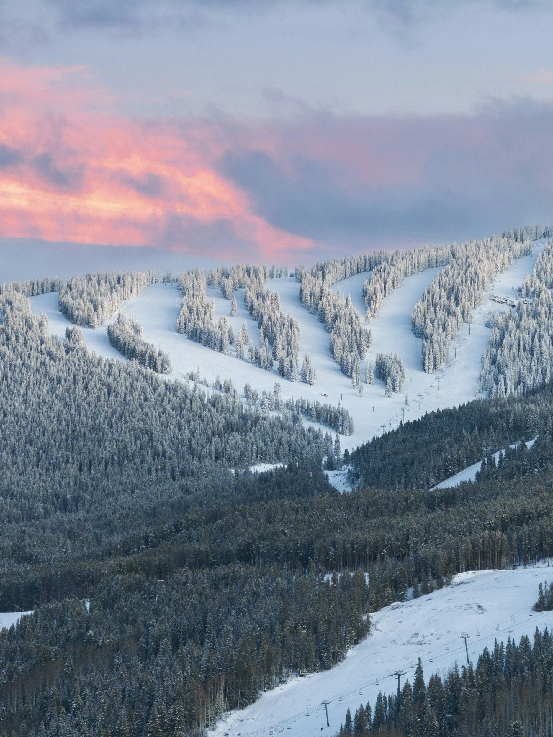 Snow-covered ski slopes with ski lifts and a forested mountain landscape under a sky with pink and purple clouds.