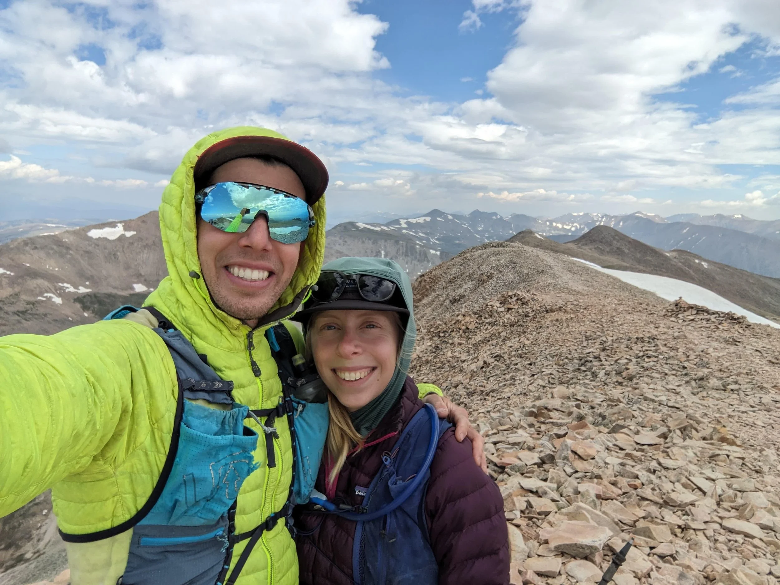 Two people taking a selfie on a mountain summit with rocky terrain and distant mountain ranges in the background. They are wearing outdoor gear and sunglasses.