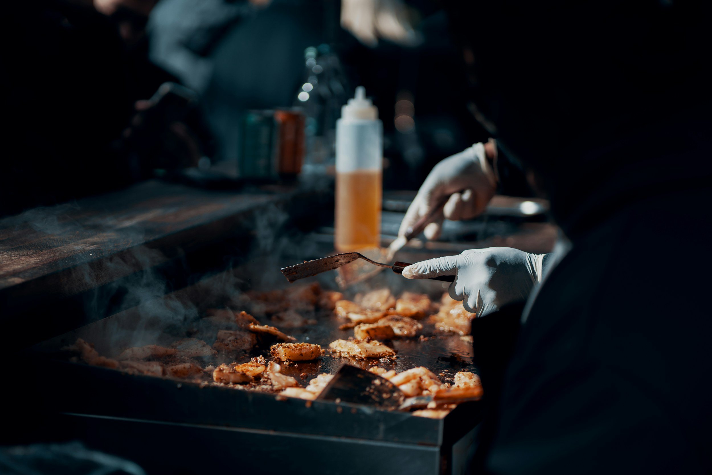 Person cooking meat on a grill, wearing gloves, with sauce bottles and spices in the background.