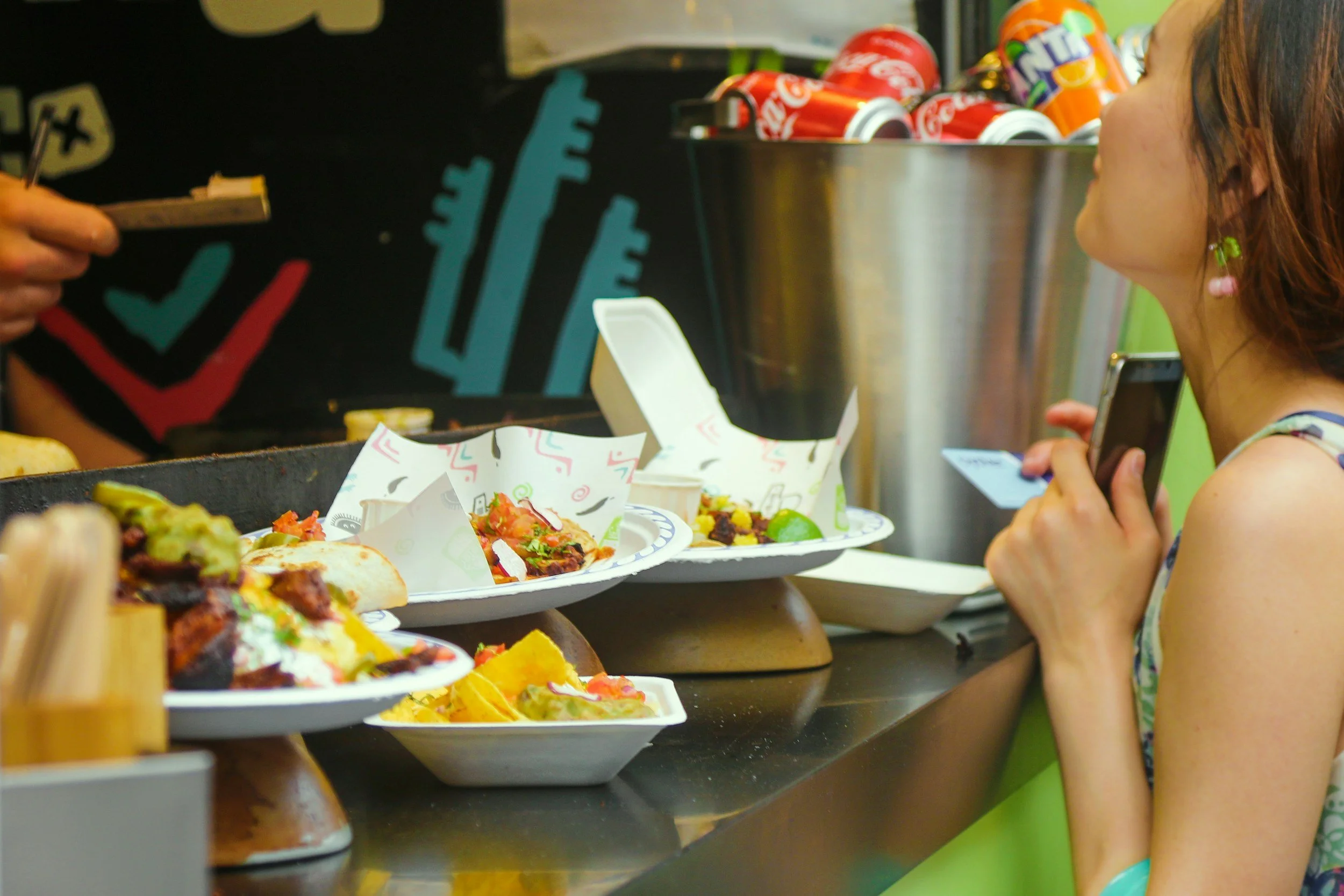 A girl with earrings holding a smartphone while ordering food at a taco stand with plates of tacos and cans of soda on the counter.
