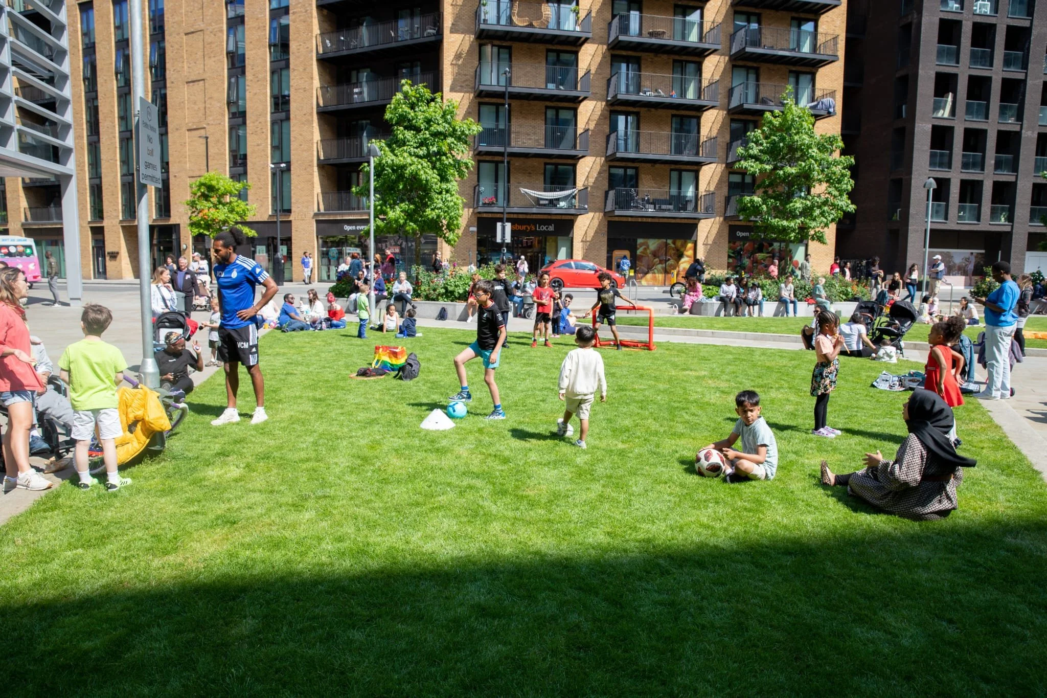 People, including children and adults, gather in a park with green grass and trees, surrounded by tall apartment buildings. Children are playing soccer and with a ball, while others sit on the grass or stand talking. Some people are watching, and others are seated in the background near the sidewalk.