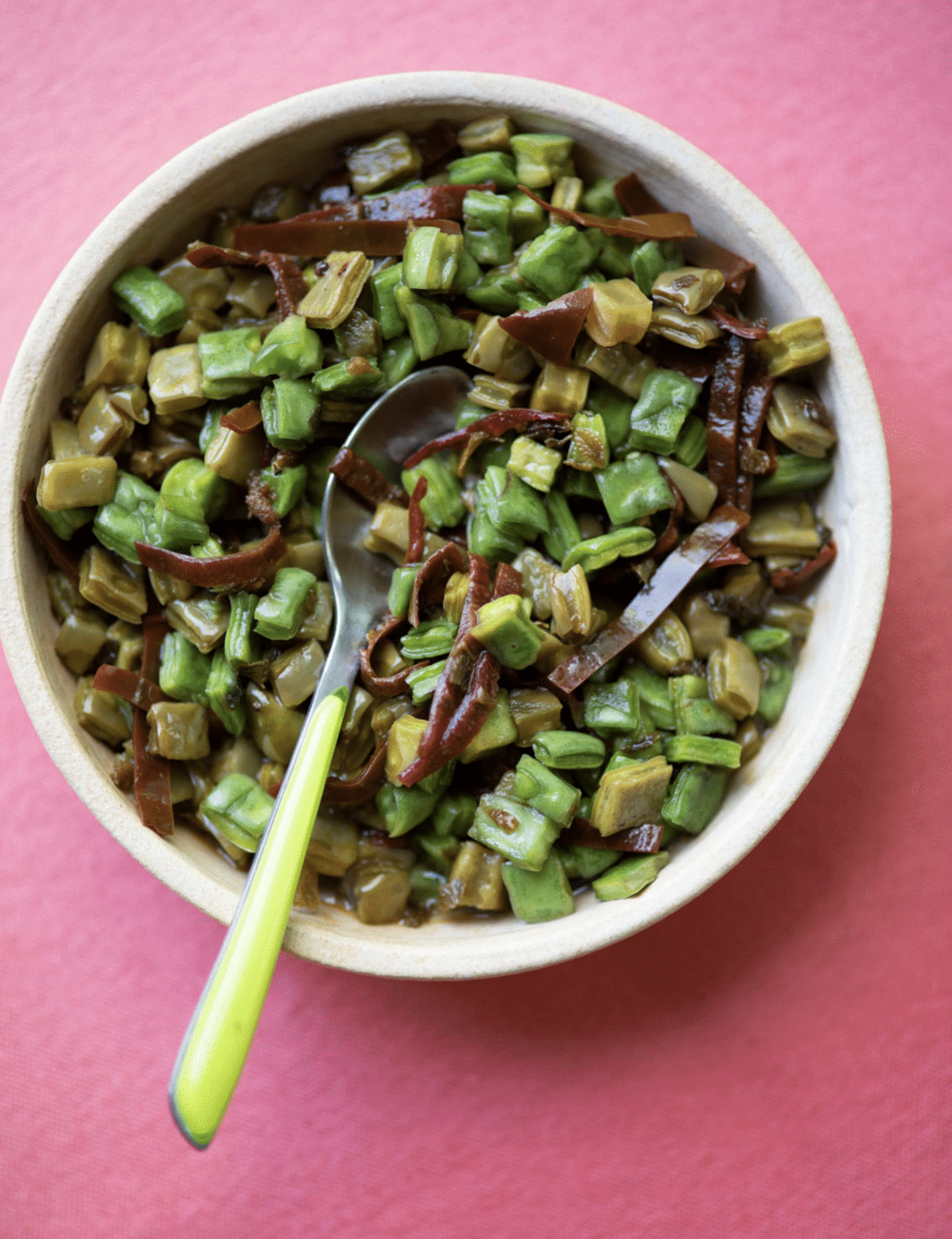 A bowl of chopped cooked vegetables with a spoon and a green straw on a pink background.