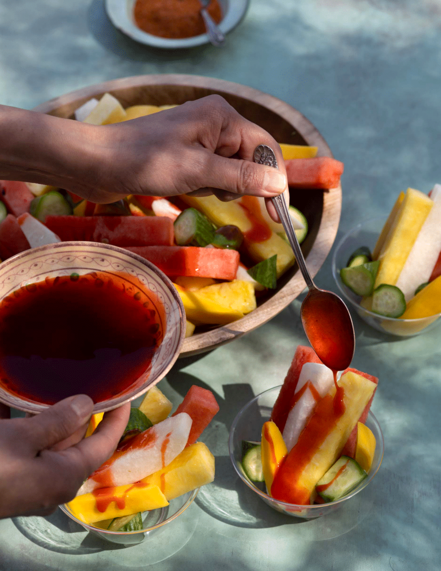 A hand holding a small spoon above a bowl of sliced watermelon, pineapple, and other fruit. In the background, there are additional bowls of fruit and sauce on a light blue surface.