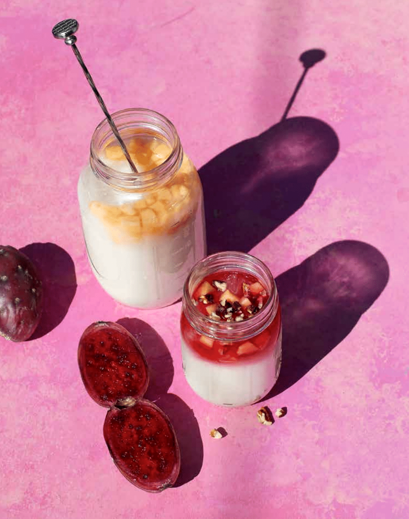 Two glass jars filled with creamy drinks and topped with fruit, set on a pink surface with strawberries and a small decorative fruit nearby, with shadows cast across the surface.