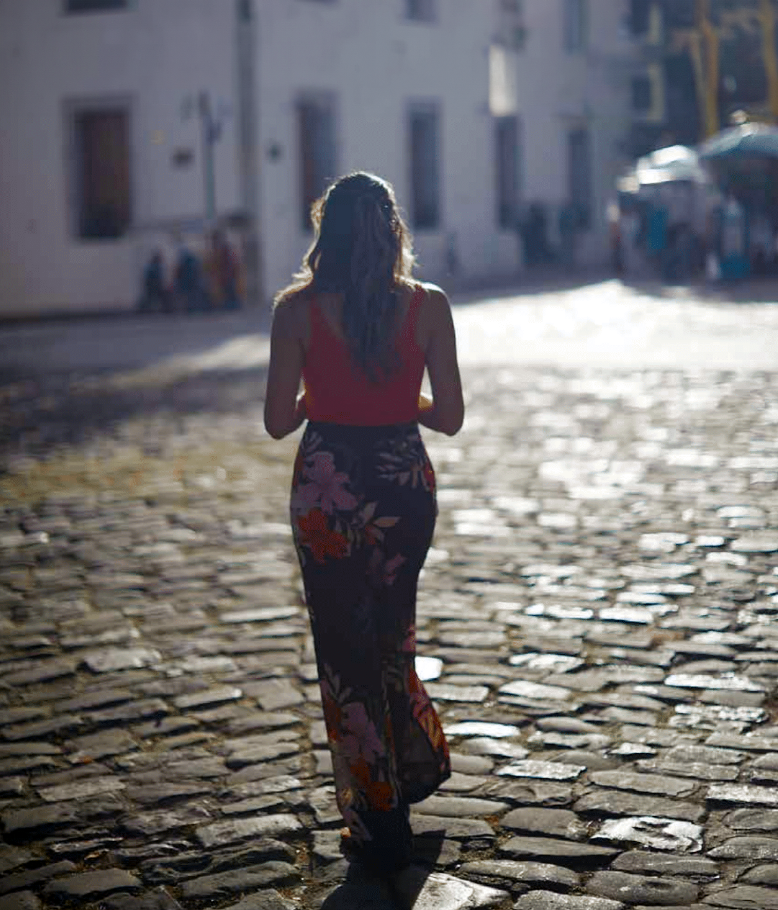 A woman with long hair wearing a red top and floral patterned pants walking on a cobblestone street in a sunny urban area.