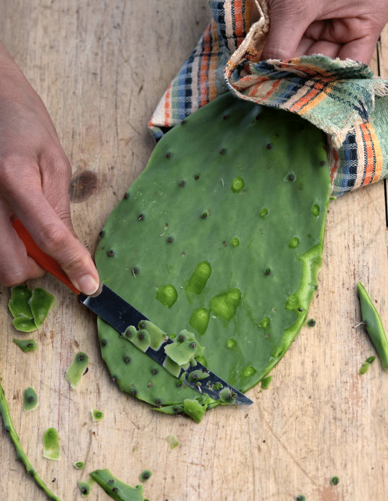 Close-up of a cactus pad being cut with a knife, with pieces and small thorns scattered on a wooden surface. A person's hand is visible holding the knife.