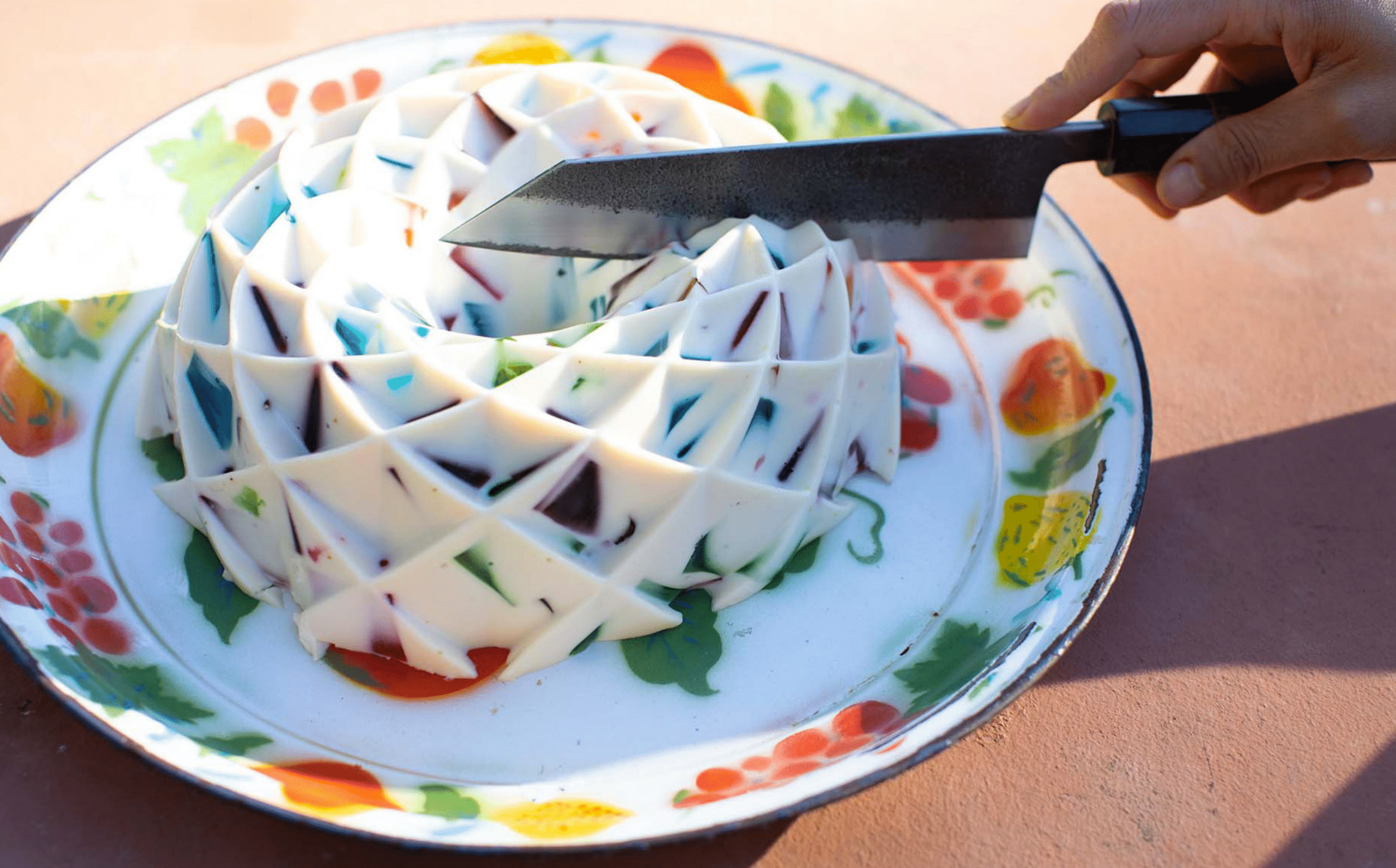 A hand slicing a circular, layered, colorful gelatin dessert on a decorative plate.