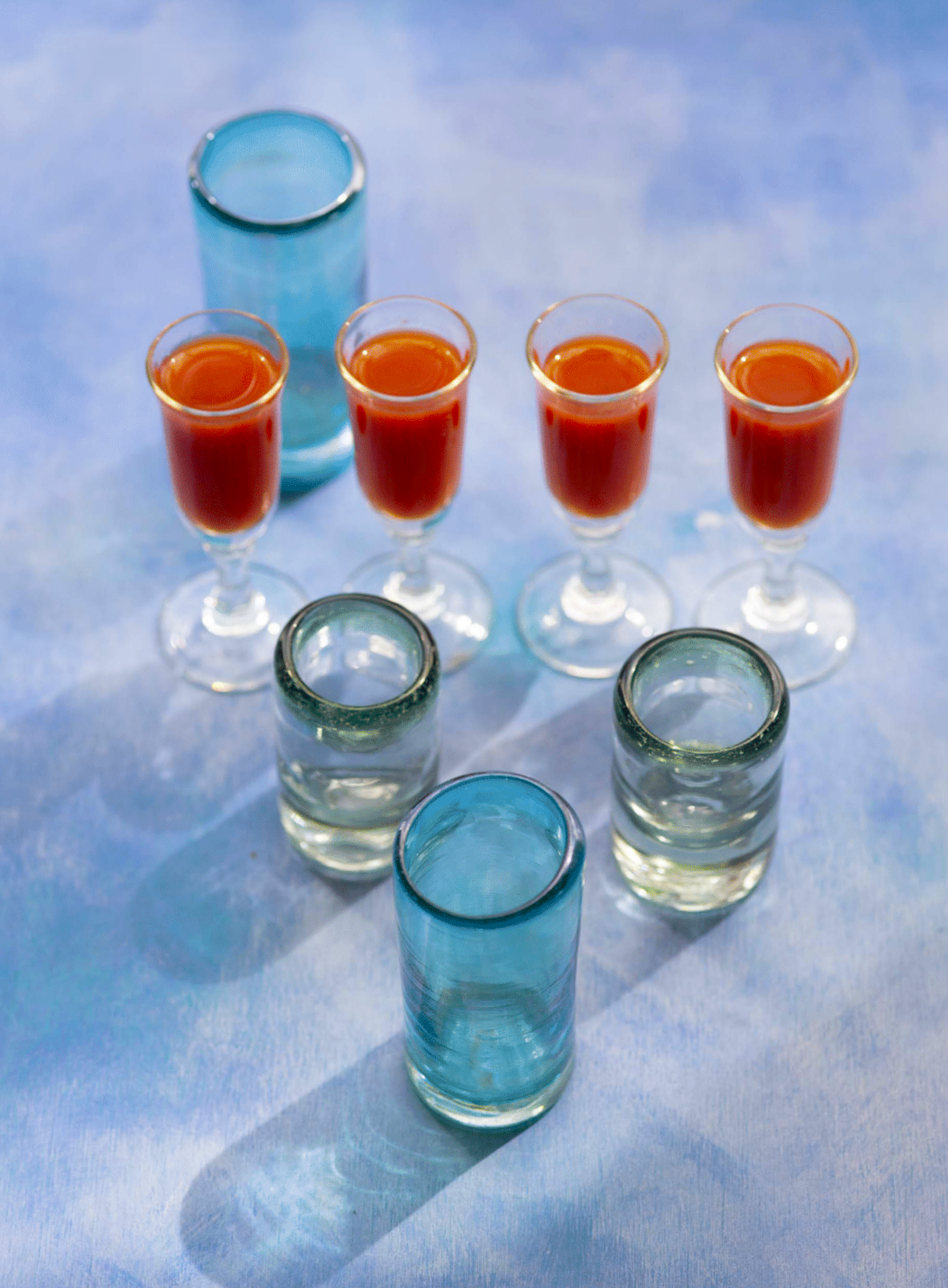Multiple transparent shot glasses with orange and clear liquids arranged on a light blue surface, with blue shot glasses in the background.