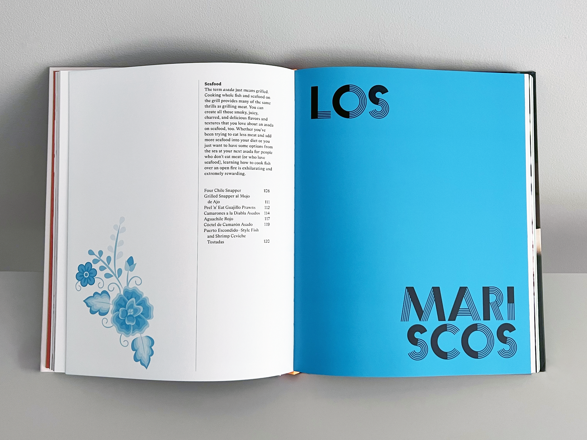 Open cookbook on gray surface showing pages about seafood, with a blue right page with black stylized text that reads "LOS MARISCOS".