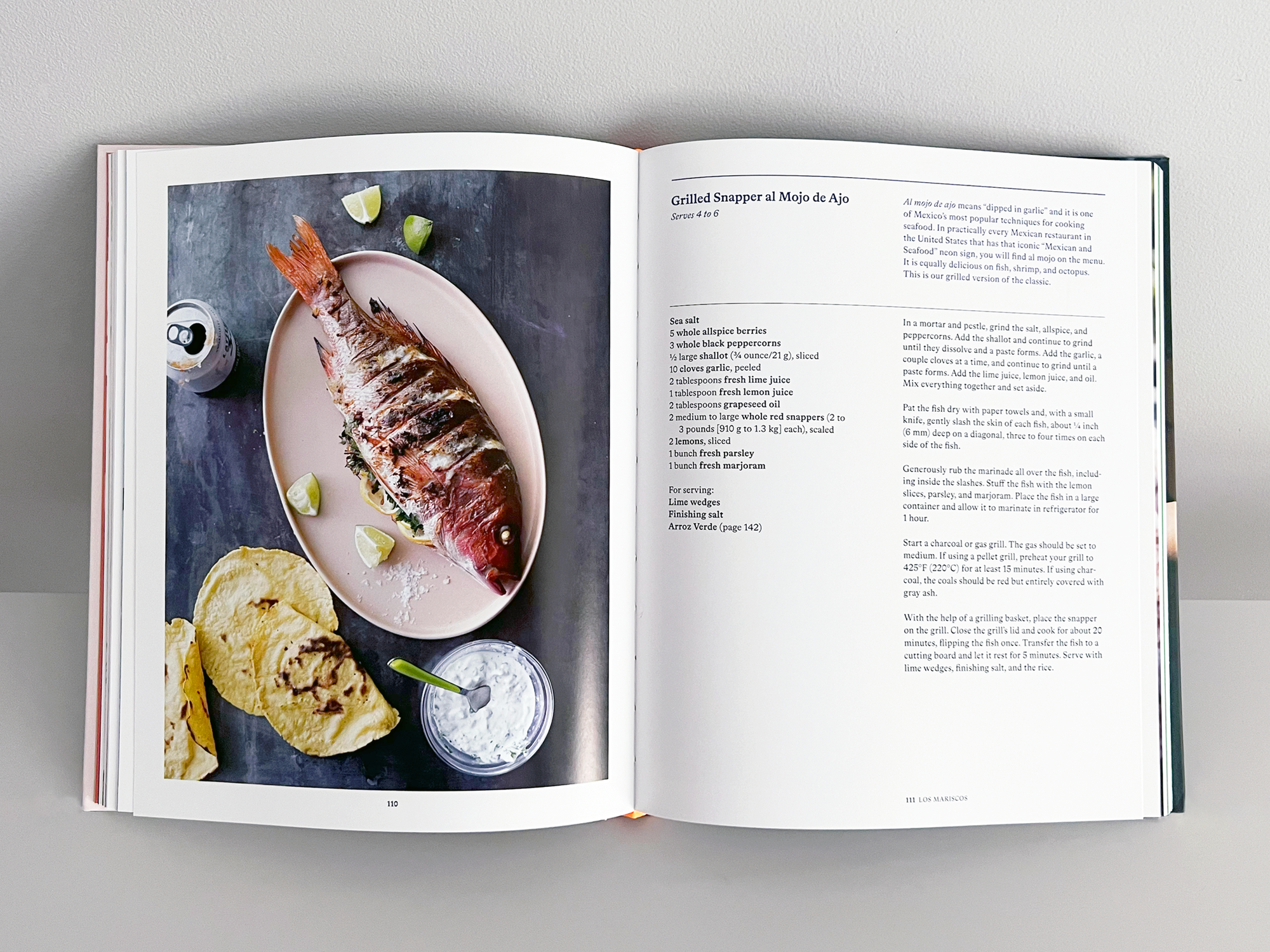 Open cookbook with a page showing a photo of grilled fish on a pink oval plate, garnished with lime wedges. The other page contains a recipe for grilled snapper al mojo de ajo, with a list of ingredients and instructions.