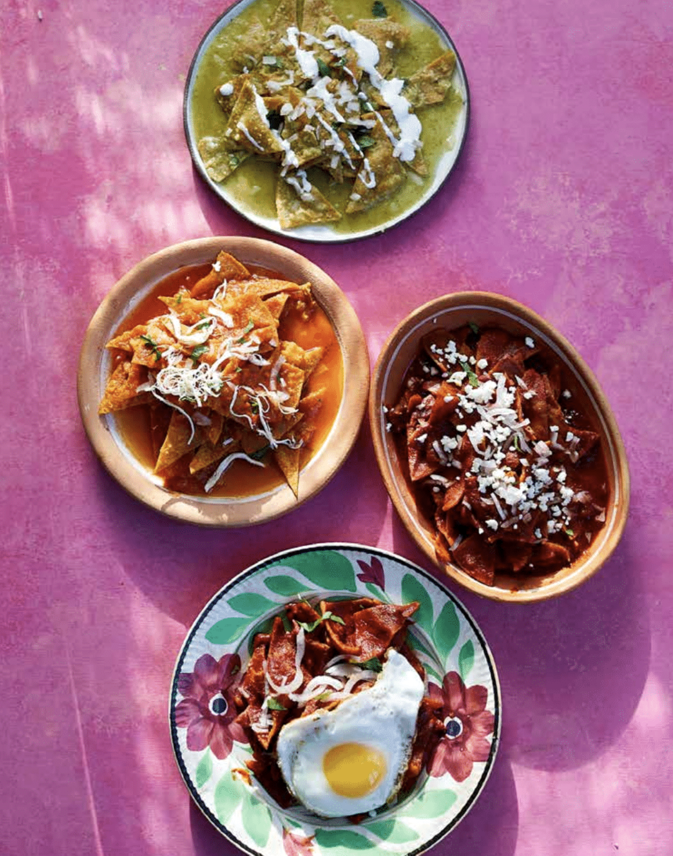 Multiple bowls of Mexican food, including tacos with various fillings, topped with cheese and garnishes. One plate includes a fried egg on top, with a pink tablecloth underneath.