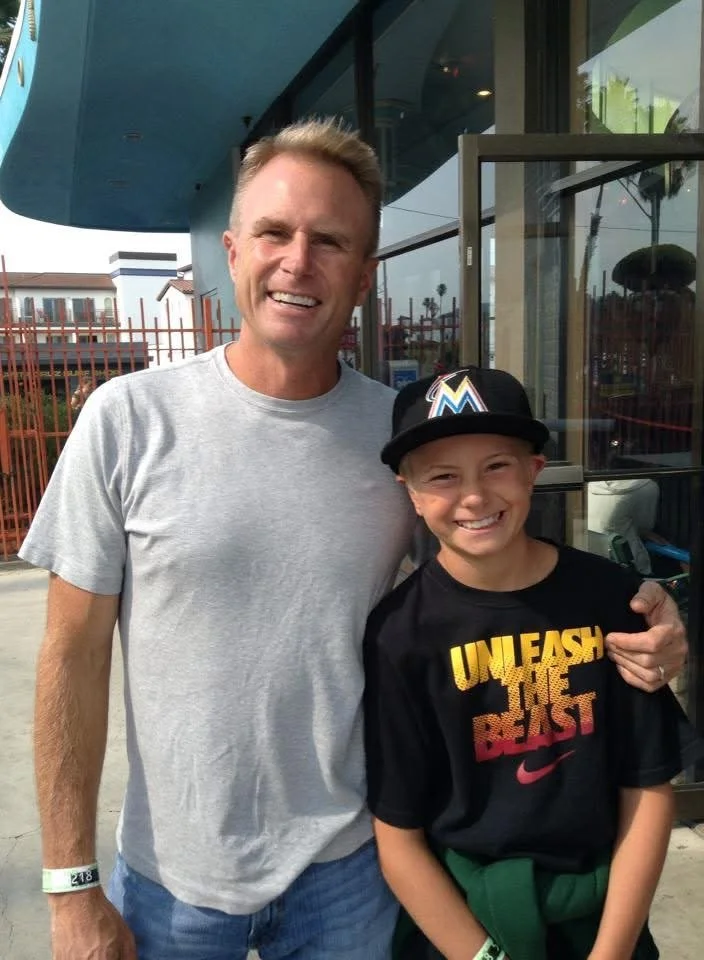 A man and a boy smiling, standing outside a building with large glass windows and a metal fence in the background. The man is wearing a light gray t-shirt and the boy is wearing a black Miami Marlins cap and a black shirt that says 'Unleash the Beast' with a Nike logo.