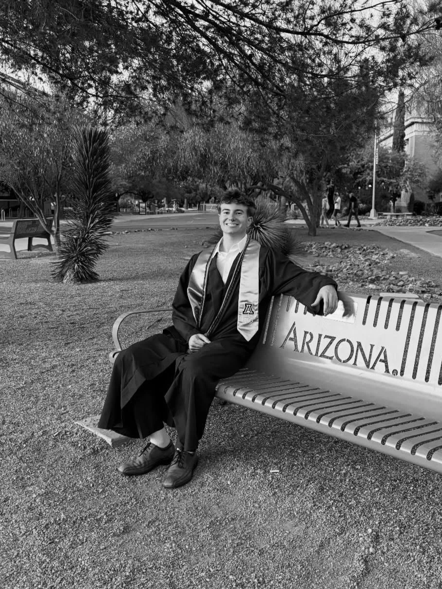 A young man sitting on a bench in a park, wearing a graduation gown and sash, smiling at the camera. The bench has the word "Arizona" inscribed on it. Trees and other people are visible in the background.