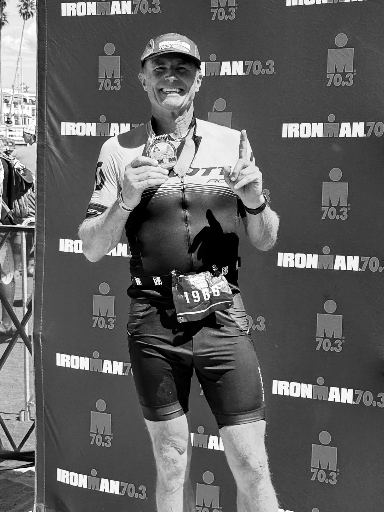 An older male athlete in running gear holding a finisher medal at the Ironman 70.3 race, smiling and pointing upward.