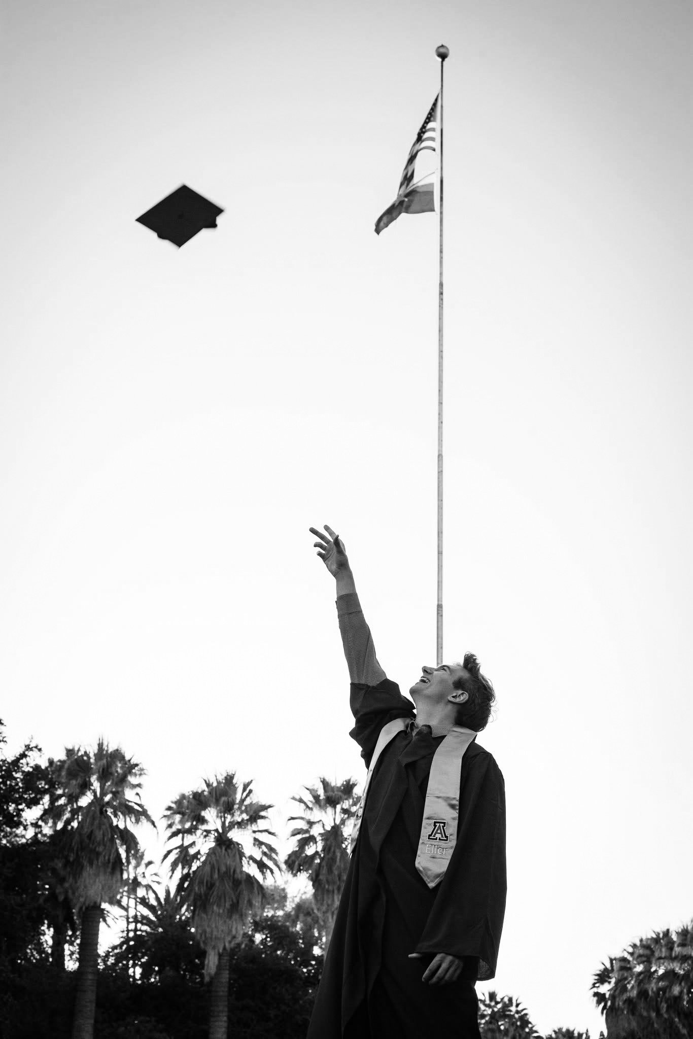 A woman in a graduation gown and sash throws her graduation cap into the air, smiling, with palm trees and an American flag flying in the background.