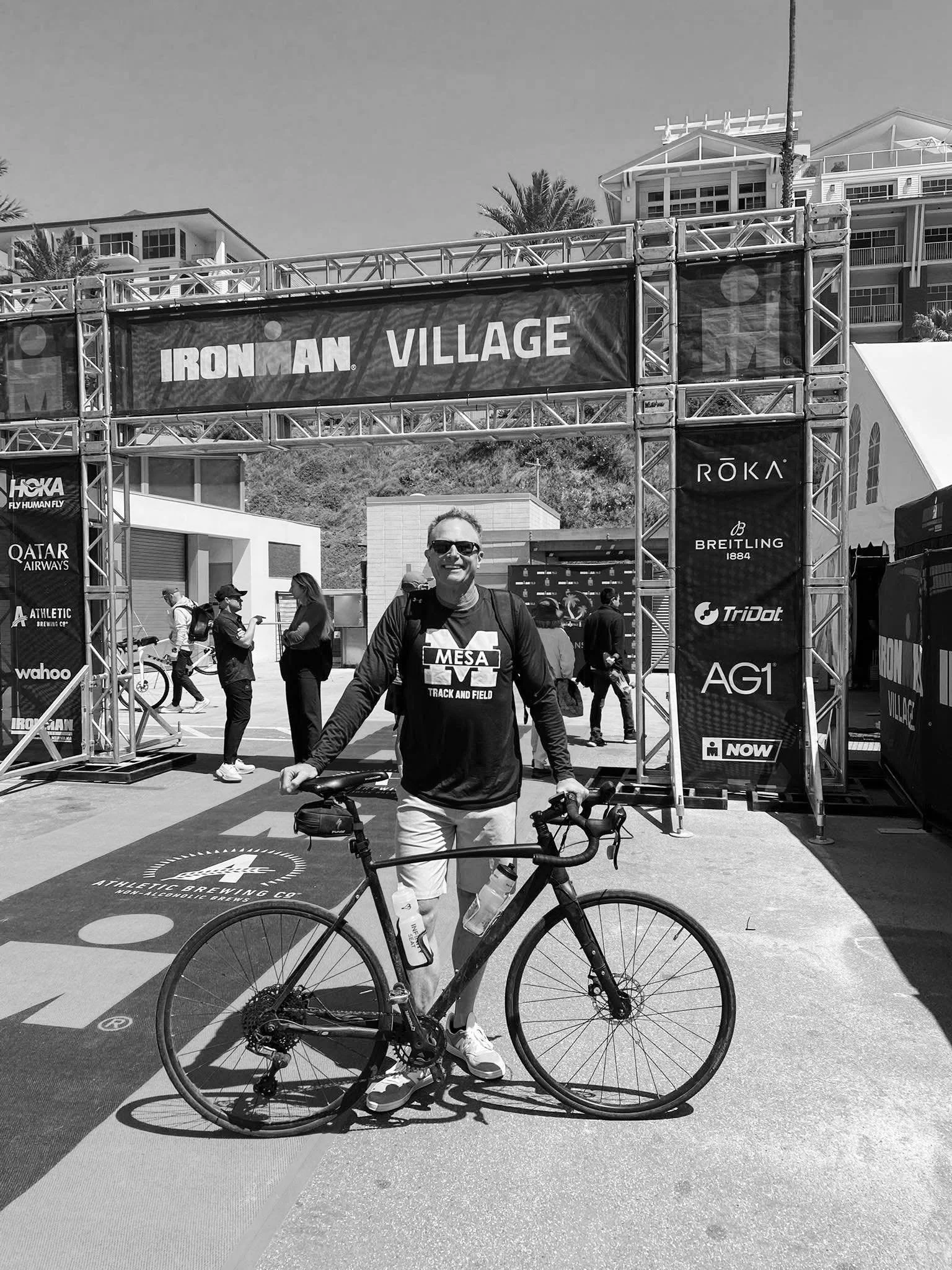 A man standing with a bicycle under an Ironman Village sign at an event, wearing a MESA Track and Field shirt, sunglasses, and smiling.