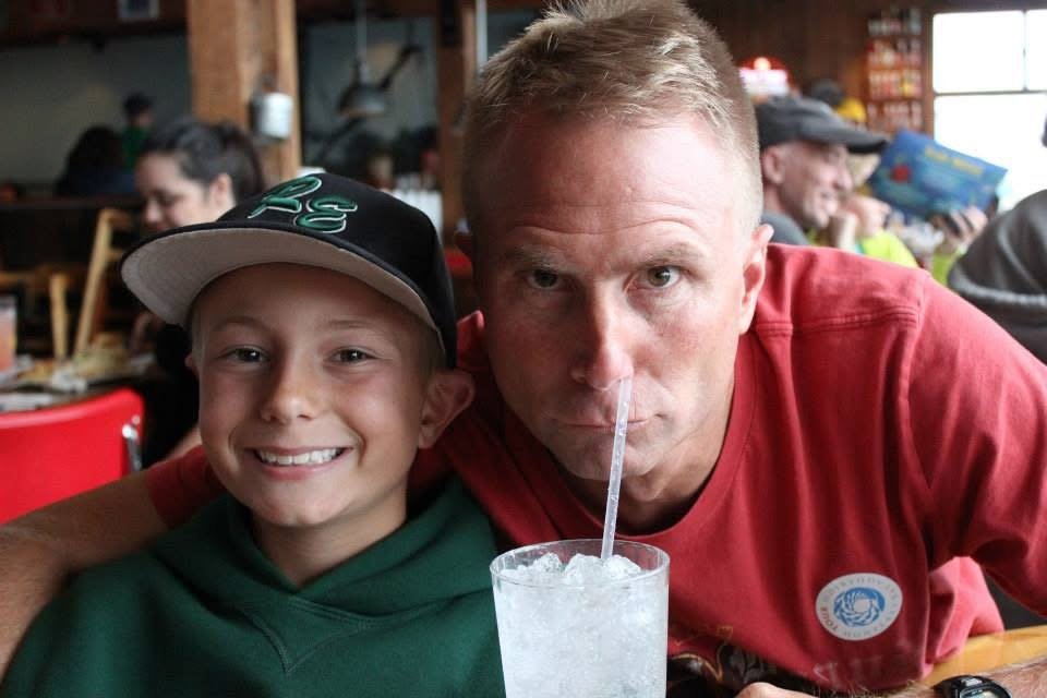 A smiling boy in a green hoodie and baseball cap sits next to a man in a red shirt drinking a beverage with a straw at a restaurant.