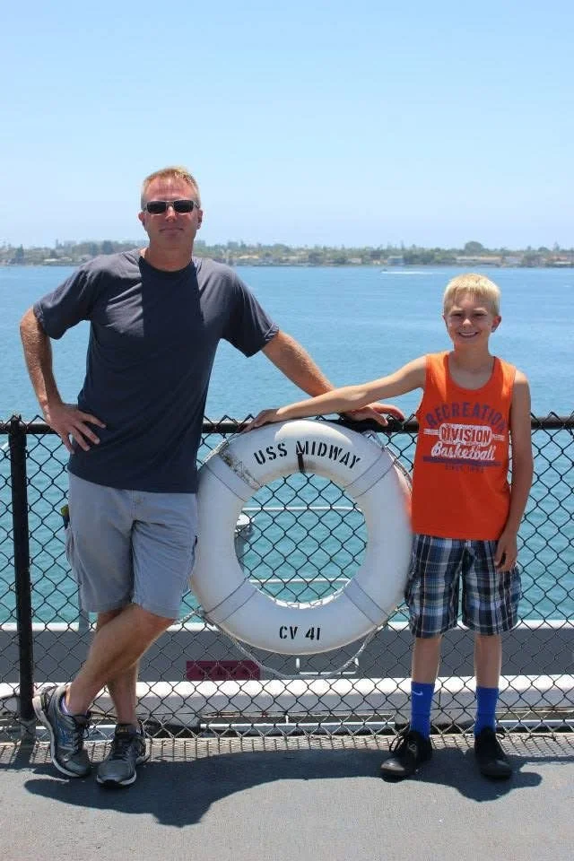 A man and a boy standing onboard a boat near a body of water, posing behind a life preserver labeled "USS Midway CV 41." The man is wearing sunglasses, a dark T-shirt, and shorts, while the boy is wearing an orange sleeveless shirt and plaid shorts. In the background, water and distant land are visible.