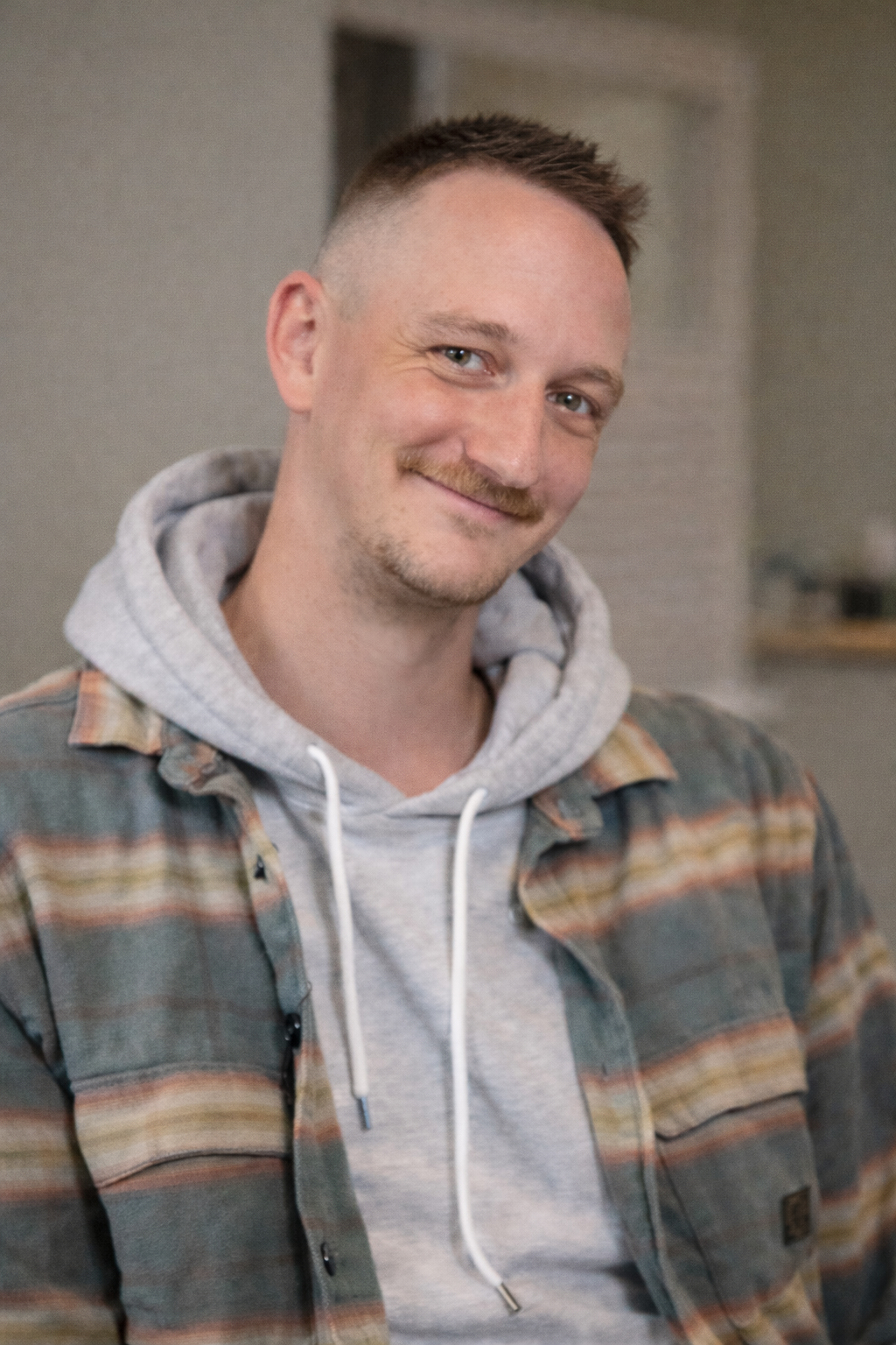 A young man with short hair, a mustache, and a beard, smiling while looking at the camera. He's wearing a gray hoodie and a plaid flannel shirt, with a blurred indoor background.
