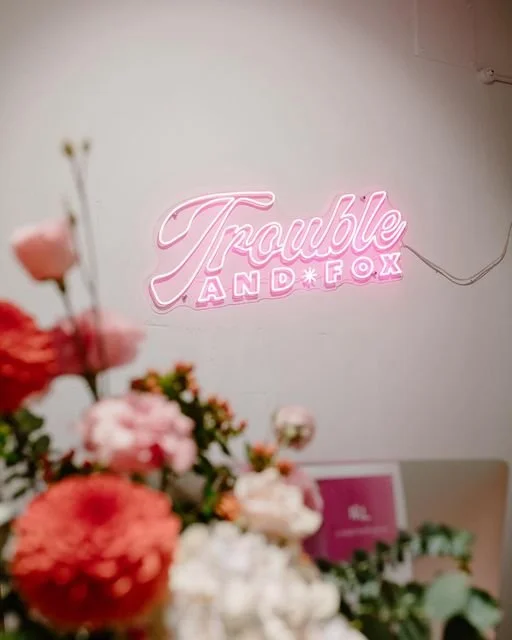 Pink neon sign that says "Trouble and Fox" on a white wall, with blurred flowers in the foreground.
