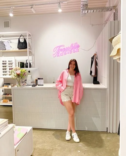 A young woman with long dark hair standing inside a boutique store, smiling, wearing a pink jacket, white top, beige shorts, and white sneakers, in front of a minimalist white counter with a neon sign that reads 'Trouble' in pink, and shelves display