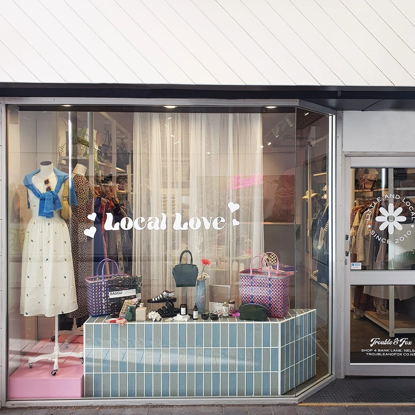 Storefront window display with clothing, handbags, and accessories, featuring a sign that says 'Local Love' with hearts, and a flower logo that reads 'Little and Local Since 2010'.
