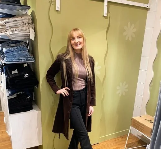 A woman with long blonde hair smiling and posing indoors against a green wall with floral patterns, surrounded by shelves of folded clothes and boxes.