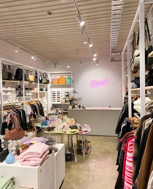 Interior of a boutique store with shelves of handbags, clothing, accessories, and a neon pink sign that says "Feminine" at the back.