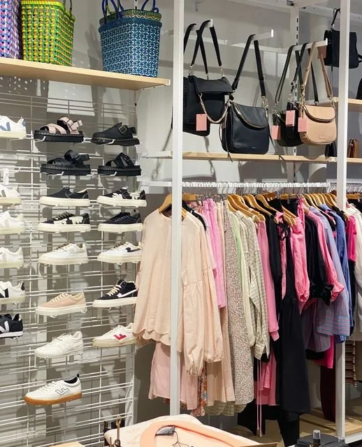 Clothing and accessories display in a retail store, featuring shoes on the left, handbags on the top shelf, and women's clothing hanging on racks below.