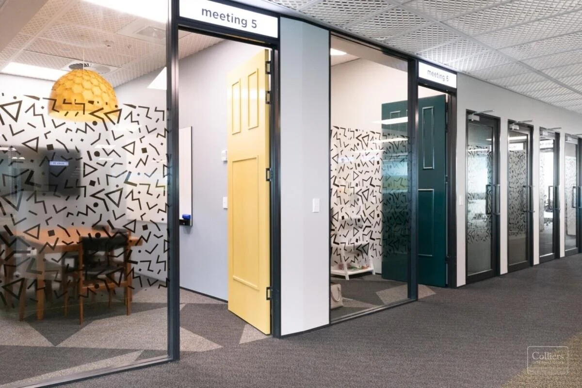 Office corridor featuring multiple glass conference rooms with black geometric patterns on the glass, labeled as meeting 5 and meeting 6, with a yellow door for meeting 5 and a teal door for meeting 6. There is a round pendant light hanging above a m