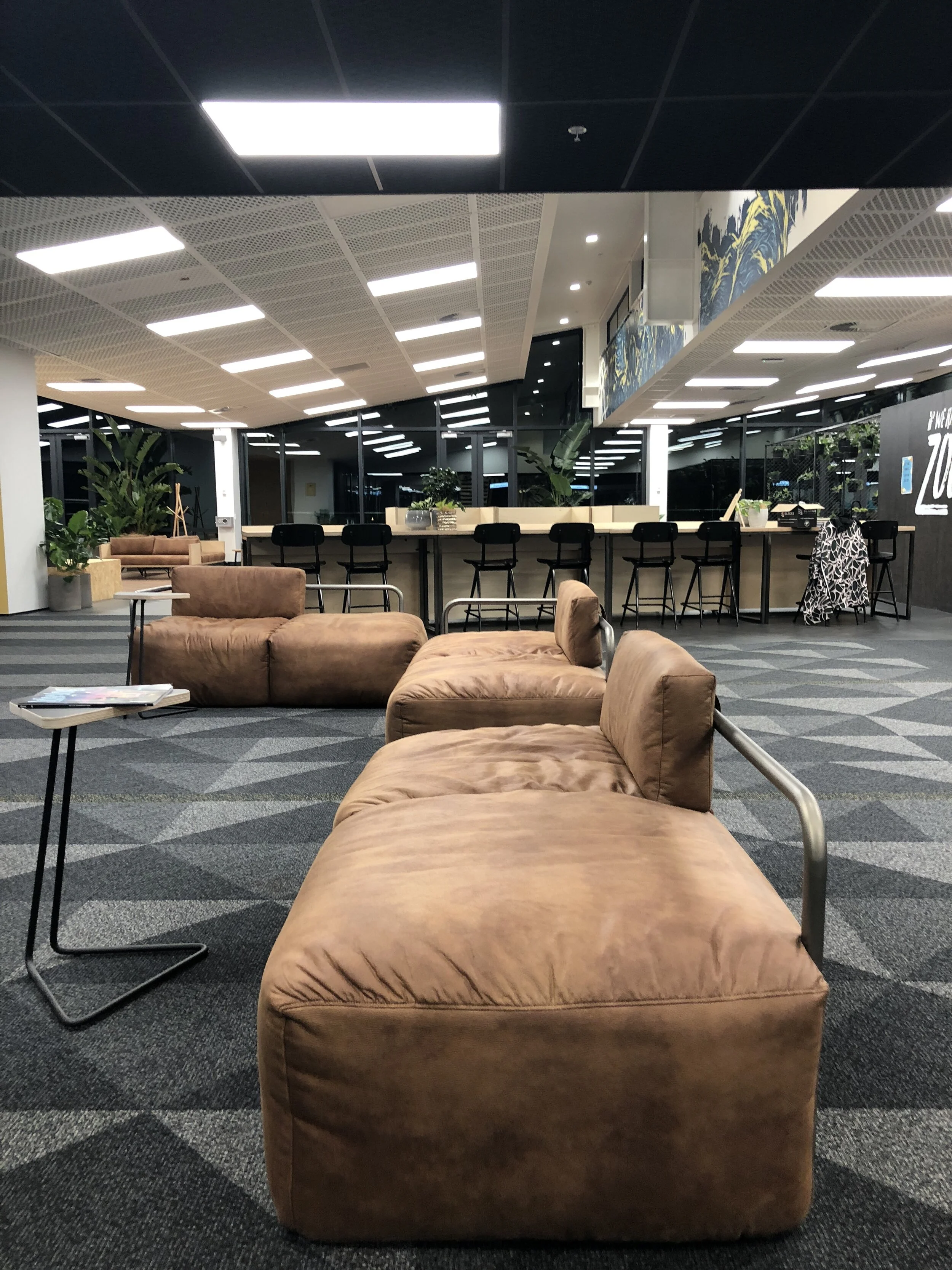 Empty airport lounge with brown sofas, a small side table, black bar stools at a counter, and potted plants near large glass windows.