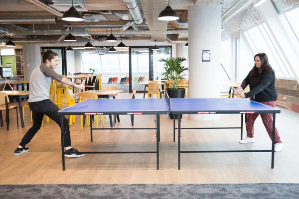 Two people playing ping pong in a modern, brightly lit office space with large windows, tables, and chairs in the background.