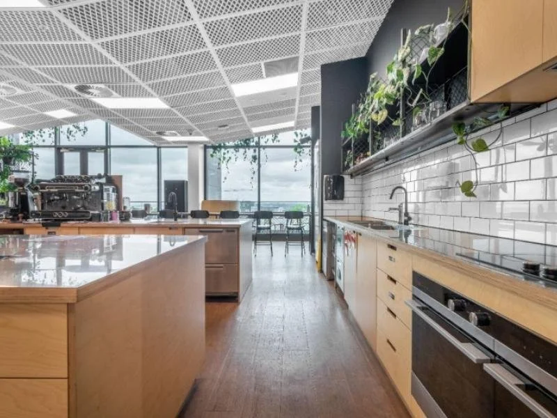 Modern kitchen with wooden cabinets, stainless steel appliances, white subway tile backsplash, and hanging plants. Large windows with city view, and an open space with tables and chairs in the background.