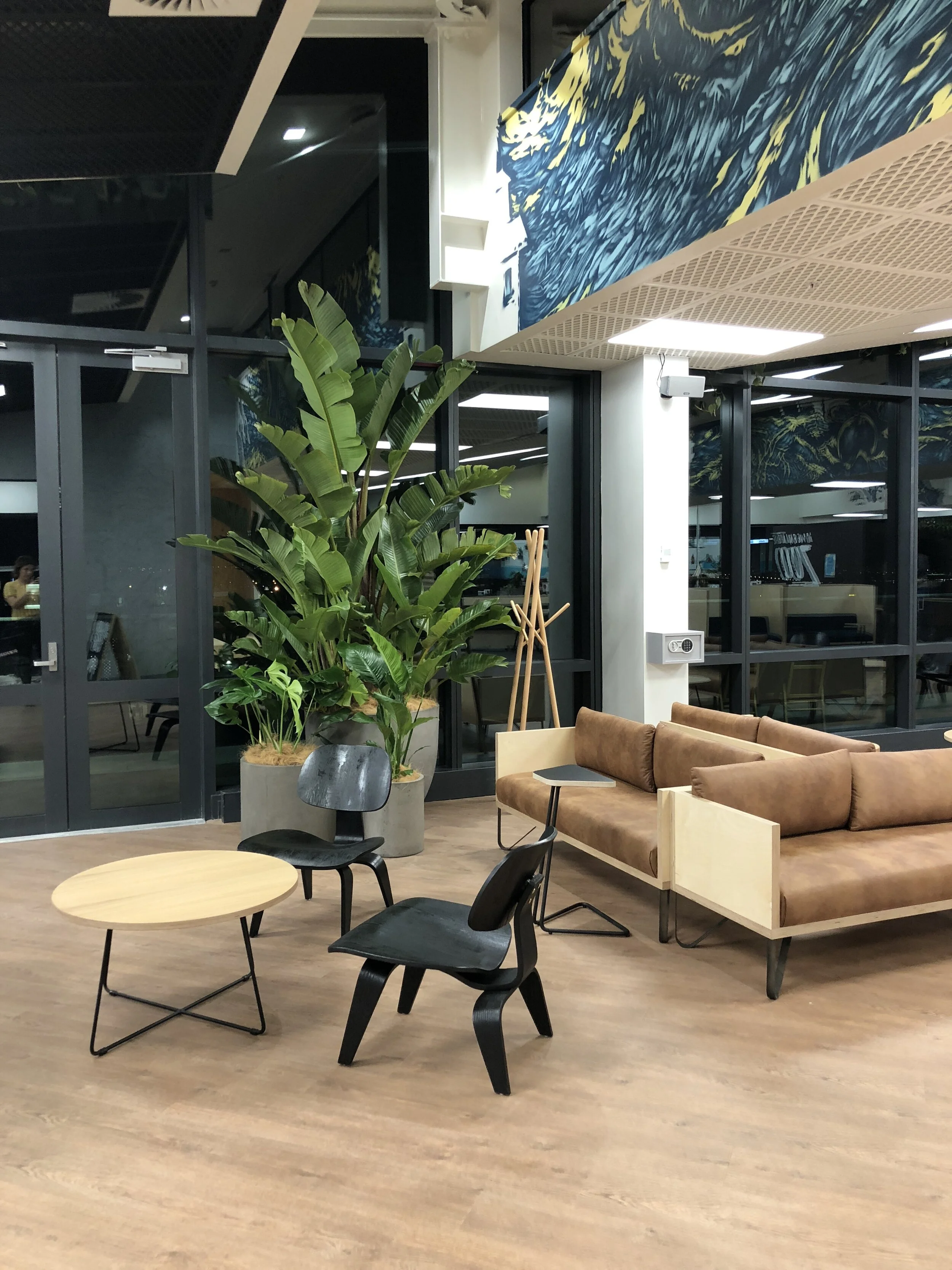 Modern lounge area with brown leather sofa, black and grey chairs, a round wooden coffee table, large green plants in concrete planters, a coat rack, and glass walls with abstract artwork above.