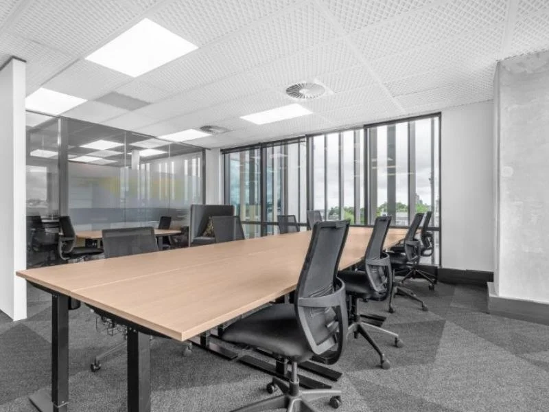Modern conference room with a long wooden table and black office chairs, large windows with blinds, and a glass-walled meeting area.