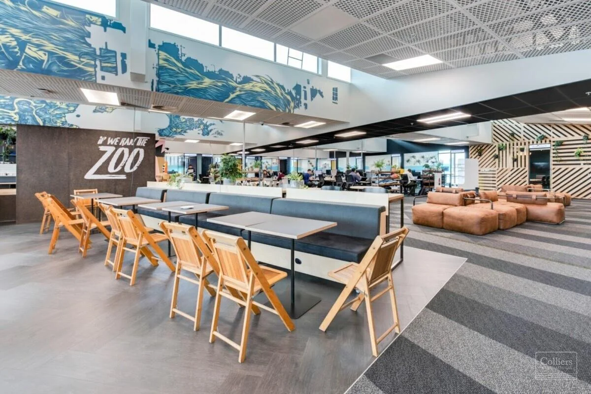 Modern office lounge with various seating options, including wooden chairs, a long bench with blue cushions, and brown leather sofas, with a large mural and greenery in the background.