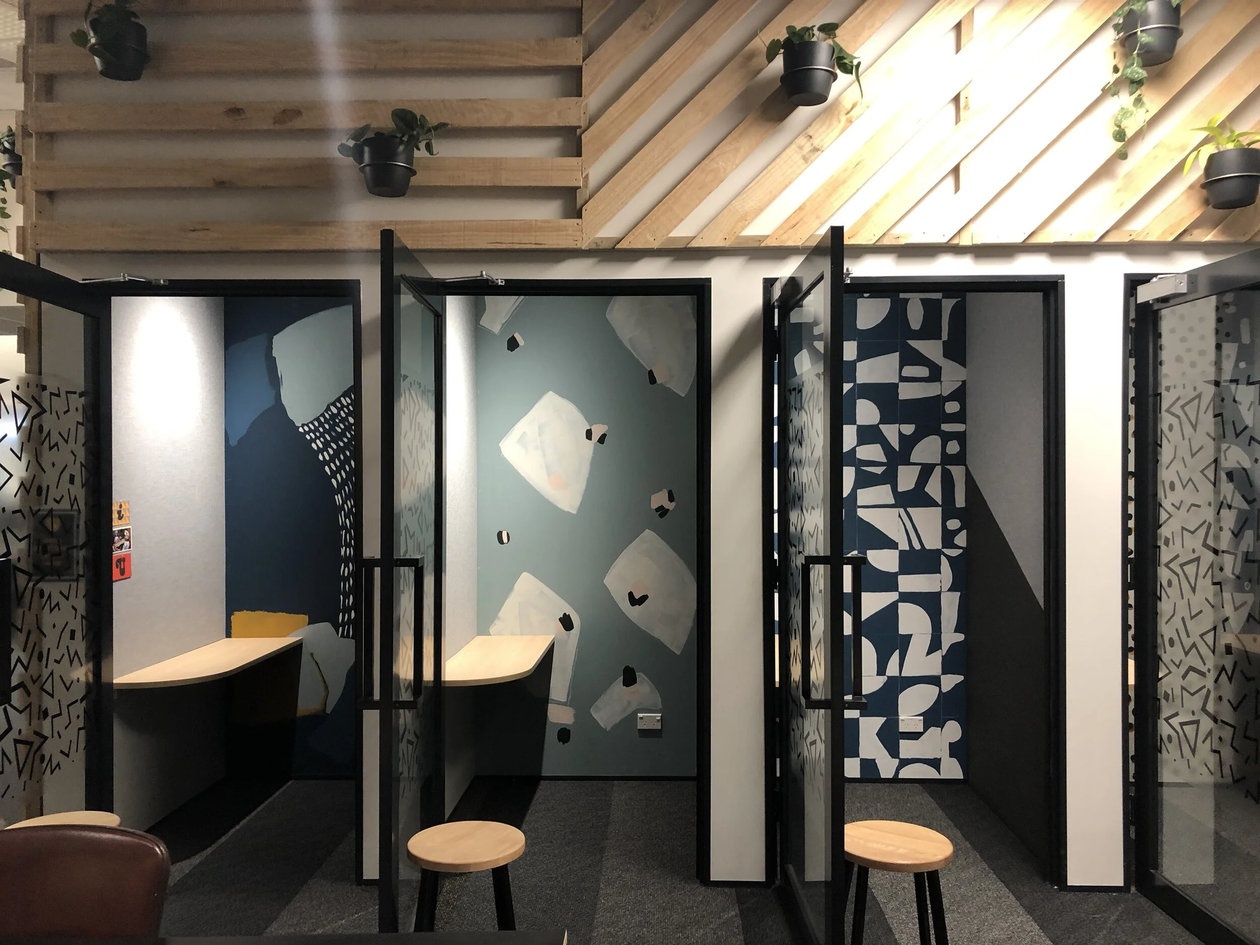 Three semi-private work booths with abstract, colorful wall murals, wooden stools, and black metal frames, inside a modern office space with a wooden slatted ceiling and hanging plants.
