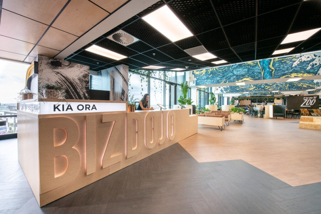 Modern lobby with a reception desk labeled 'BIZ DOJO' and the Kia Ora sign, seating area with beige sofas, large windows with plants, and decorative ceiling panels with blue and yellow patterns.