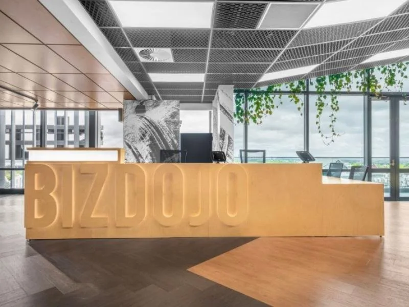 Modern office reception area with a large illuminated sign spelling BIZDOJO in front of a reception desk, large windows with a view outside, ceiling with grid pattern, and indoor plants hanging from above.