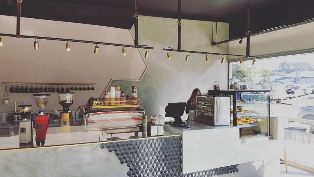 Interior of a modern coffee shop with an espresso machine, coffee grinders, and a barista working behind the counter. Large window showing parked cars outside.