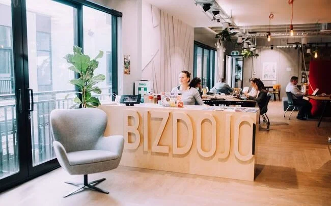 Modern coworking space with a reception desk labeled 'BIZDOJO,' a woman working behind the desk, and people working on laptops at tables in a bright, open room with large windows and greenery.