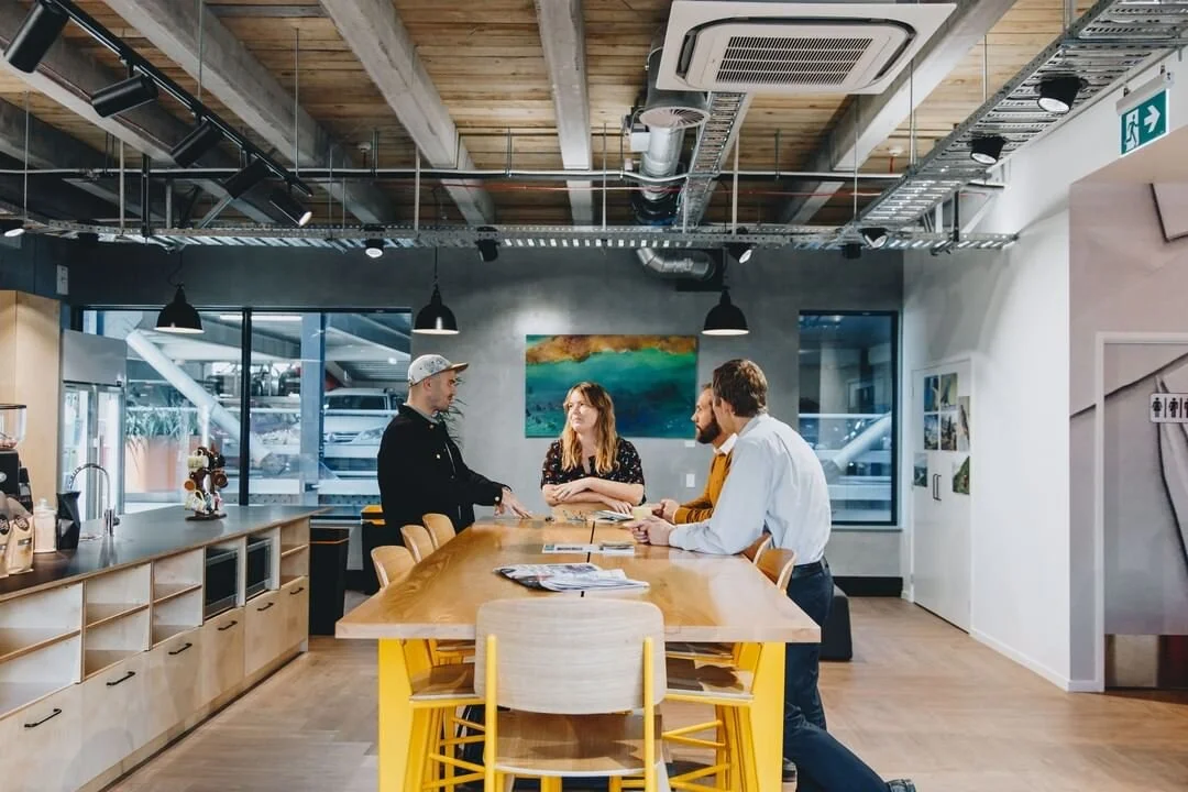 Three people are engaged in conversation around a wooden table inside a modern, well-lit cafe, with a woman and two men standing on opposite sides of the table.