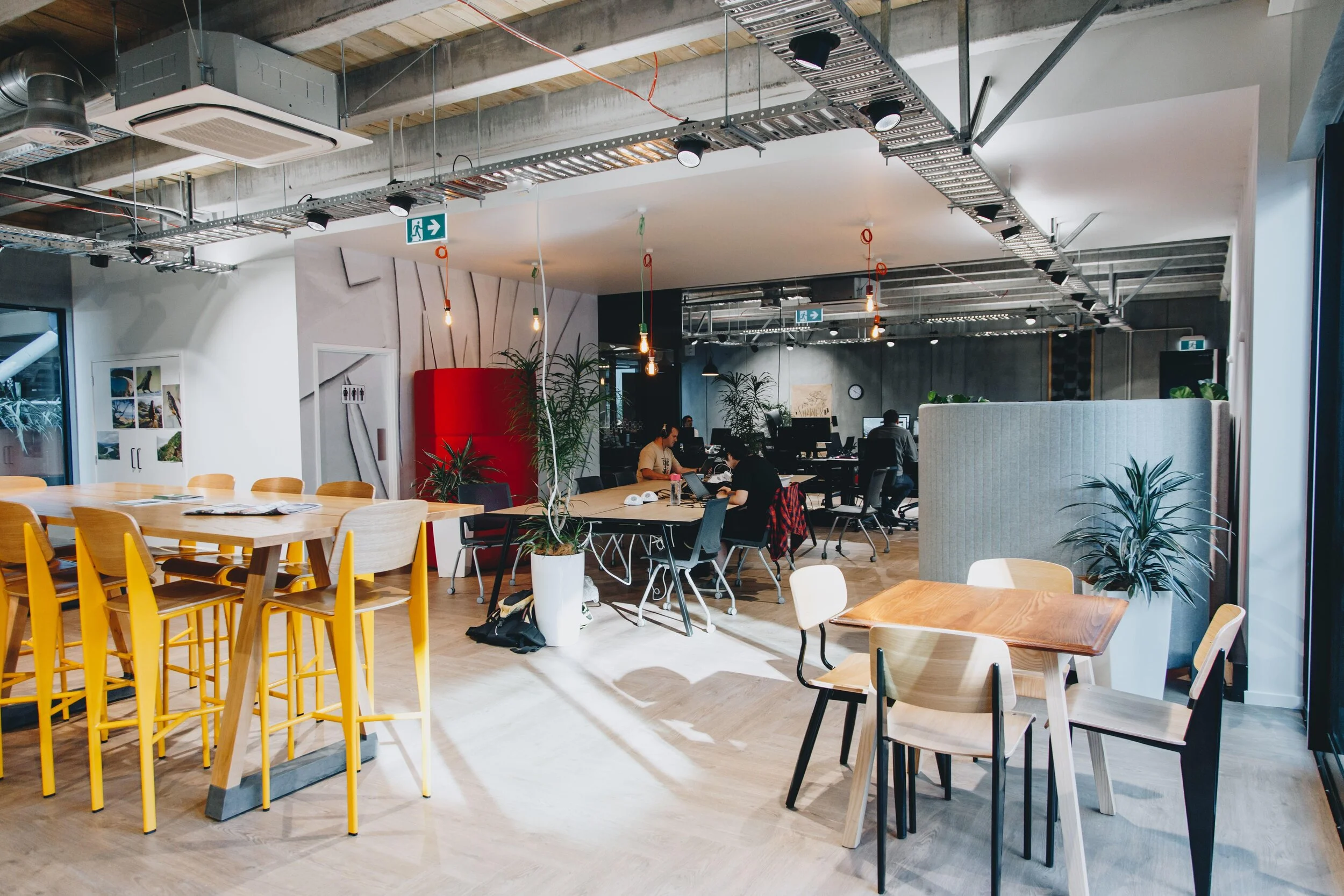 Modern coworking space with wooden tables, chairs, indoor plants, and people working at desks.