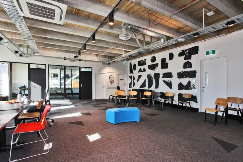 Modern office lounge with colorful chairs, a blue ottoman, abstract black and white wall art, and large windows letting in natural light.