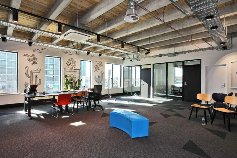 Modern office space with large windows, wall art, a table with chairs, a blue bench, and additional chairs near the wall, with an open ceiling showing ducts and beams.