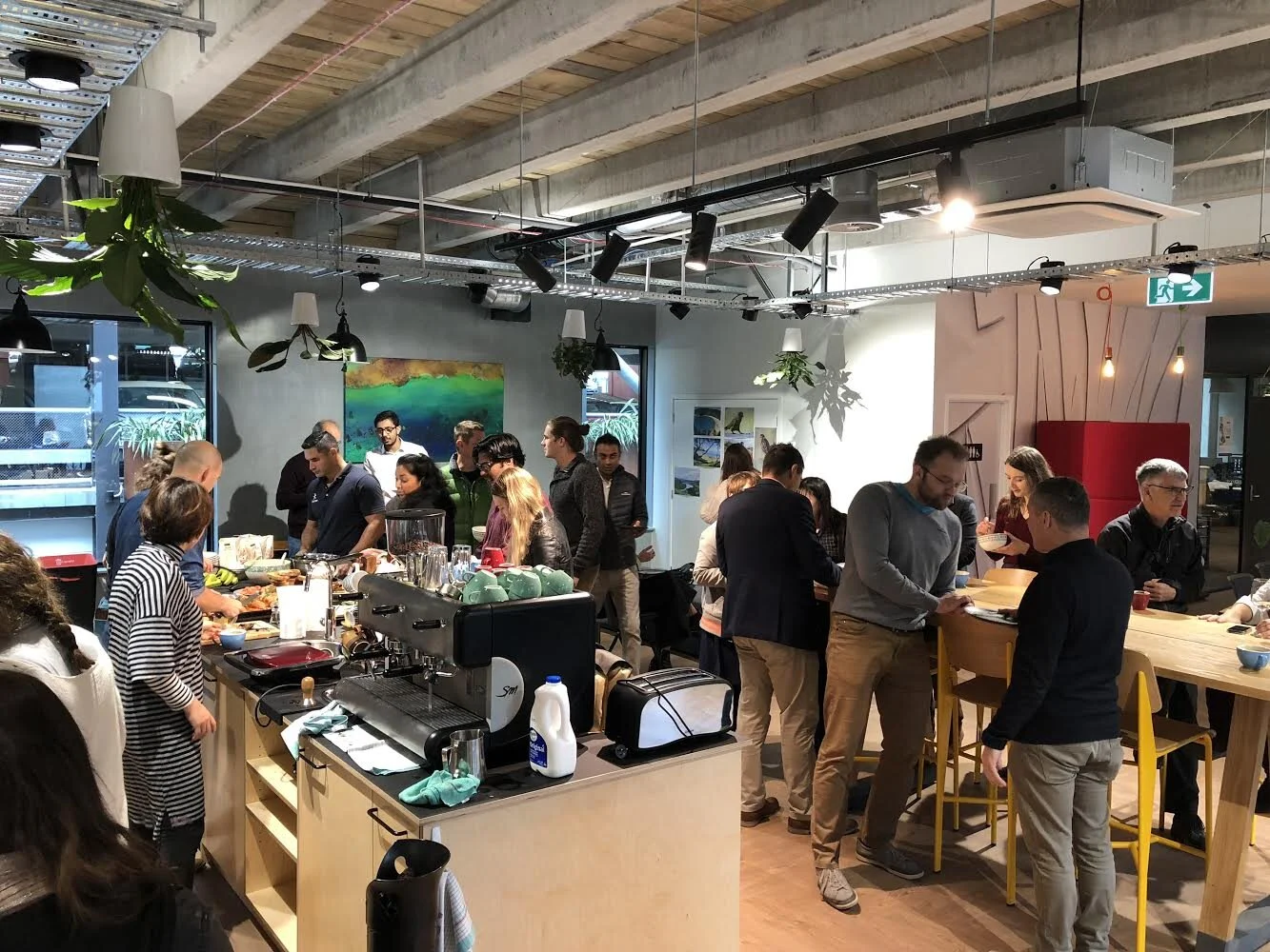 People gathered in a modern, well-lit cafe or restaurant with a buffet-style setup, coffee machine, and bar-height tables. Some individuals are serving themselves food, while others are chatting or reading menus. The interior features hanging plants,